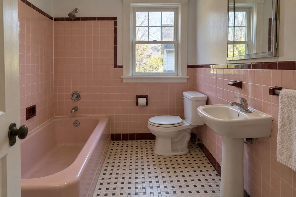 Dated 1950s bathroom with pink wall tile, a maroon trim row, a pink built-in tub and worn floor tile before a remodel